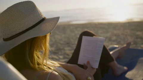 Beautiful woman sitting at the beach at sunset, reading a book Stock Footage 59336049