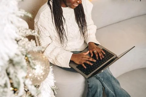 Beautiful young african female sitting with laptop near Christmas tree at home Stock Photos