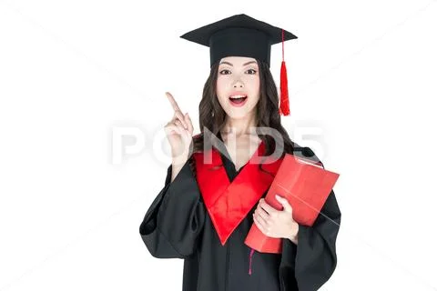 Beautiful young brunette woman in mortarboard holding book and pointing away Stock Photos