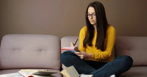 Beautiful young woman in glasses sitting on the sofa, noting in notebook Stock Footage