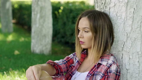Beautiful young woman sitting under the tree on the green grass at city park. 4K Video stock 88334341