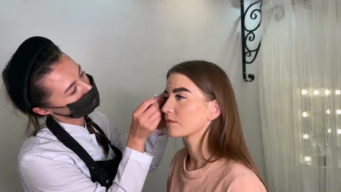 Beauty master in a mask draws a contour of eyebrows on a young girl Stock Footage 200481253