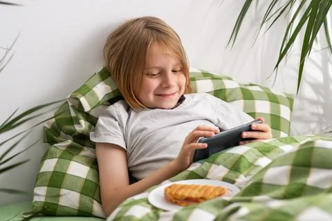 In the bed, a 9-10-year-old boy with lengthy hair savors his time online, e.. Stock Photos
