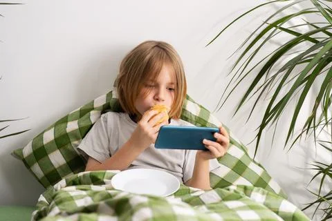 In the bed, a 9-10-year-old boy with flowing locks enjoys a toast, leisurel.. Stock Photos