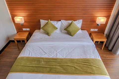 A bed with white sheets and green pillows sits against a wooden panel wall,.. Stock Photos