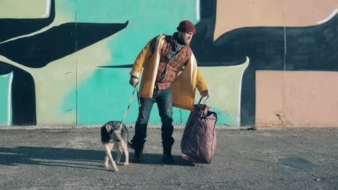 A beggar is standing next to a graffiti wall with his dog Vidéo 168129478