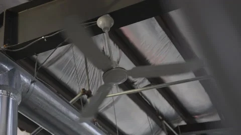 From Behind Wall Ceiling Fan Turns Next to Pipe in Warehouse Stock Footage 208756327
