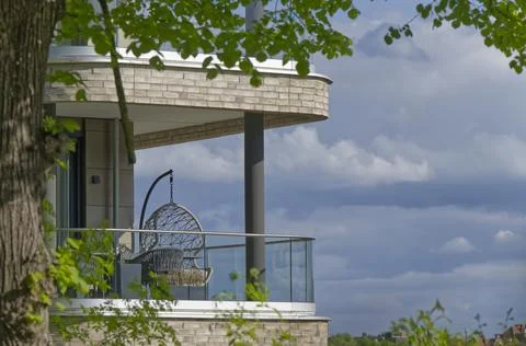 Between trees on a balcony hangs a modern empty hanging chair against a sky.. Stock Photos