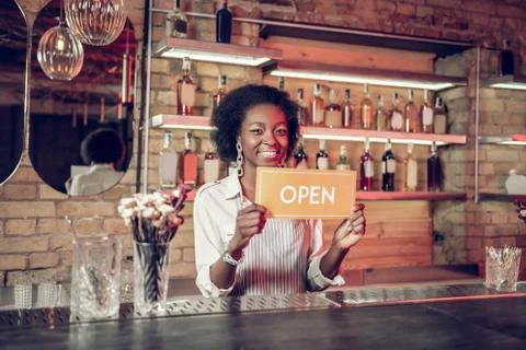 Bewitching beaming young-adult Afro-American bartender holding open sign in Stock Photos