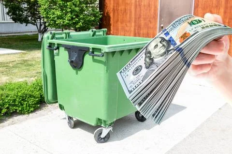 Big green plastic garbage container and stack of american dollars in the hand 스톡 사진