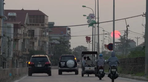 Big large orange sun sets while people drive scooters and cars down road Stock Footage 52078510