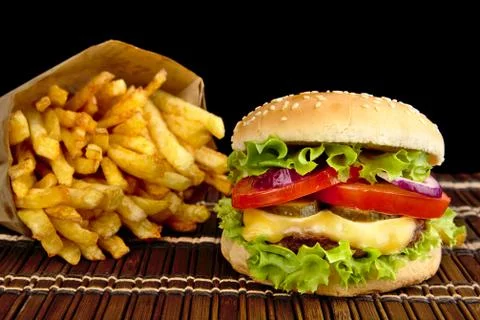 Big single cheeseburger with french fries on wooden mat on black background Stock Photos