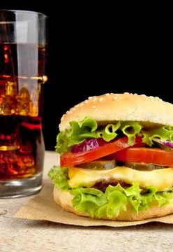 Big single cheeseburger with glass of cola on wooden desk on black background Stock Photos