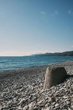 Big stone surrounded by pebbles on seashore with blue water and distant mount Stock Photos