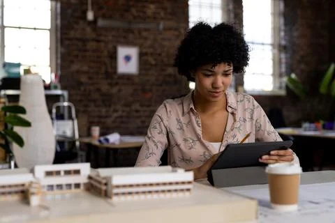 Biracial female architect using tablet sitting at desk with building model in Foto stock