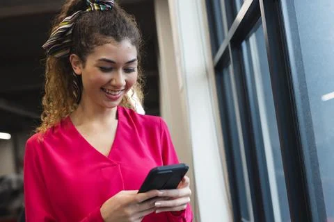 Biracial woman smiling while using a smartphone near a window at office Stock Photos
