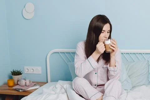 Biracial young woman in pajamas sit on bed, drink coffee cappuccino in bedroom Stock Photos