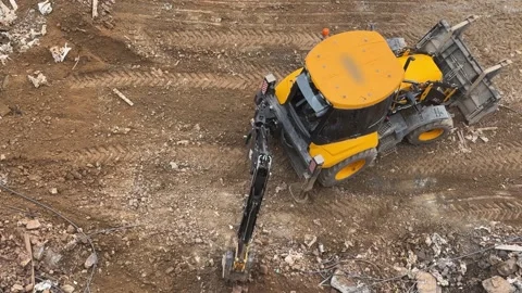 Bird's eye view of a yellow backhoe loader maneuvering on uneven and muddy Stock Footage 332395649