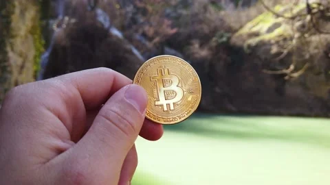 BITCOIN HELD IN HAND WITH NATURE WATERFALL IN THE BACKGROUND. UHD, 4K, Cinematic Stock Footage 150509360