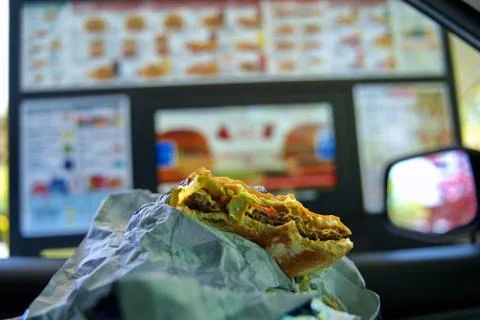 A bitten cheeseburger in the hands of a hungry driver parked by a drive up di Stock Photos