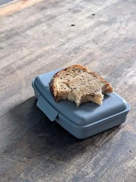 Bitten slice of rustic bread resting on closed gray lunch box over wooden tab Fotos Stock