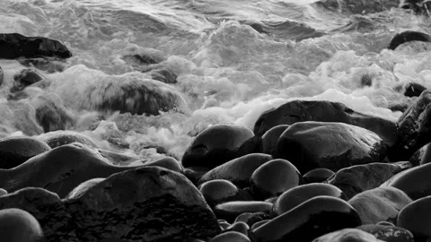 Black and white ocean waves wash over smooth dark volcanic stones on a rugged Stock Footage 346649091