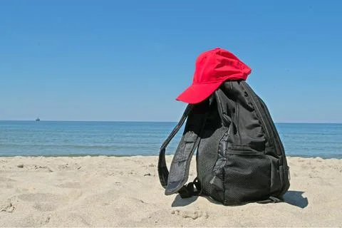Black backpack and red cap on shore of calm sea of loosened by feet gray sand. Stock Photos