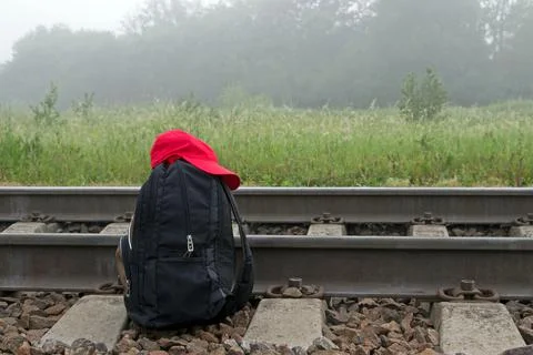 Black backpack, red cap lies next to railroad tracks on background of meadow. Stock-Fotos