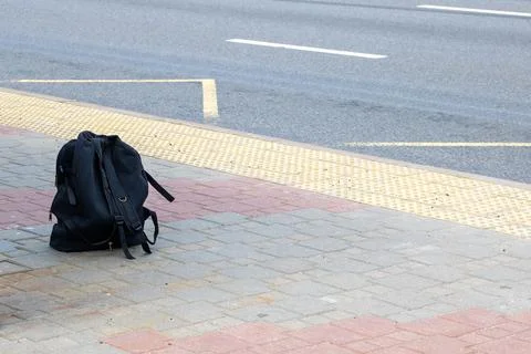 A black backpack is resting on the sidewalk that is adjacent to the road Stock Photos