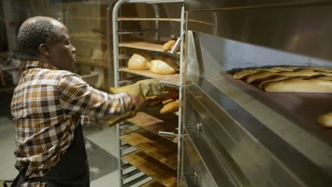 Black Baker in Gloves Removing Hot Baguettes from Oven in Bakery Kitchen Stock Footage 303462238