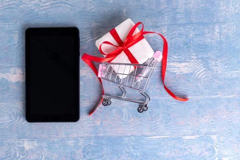 Black blank tablet or cell phone screen and trolley with gift box on blue woo Stock Photos