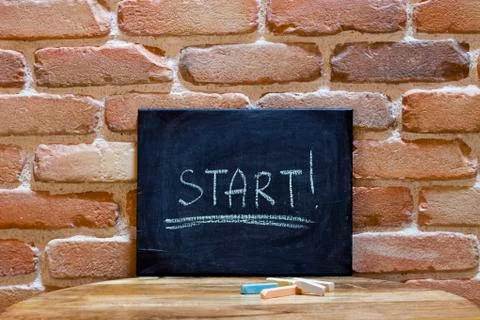 Black board with the word "Start!" drown by hand on wooden table on brick wal Stock Photos