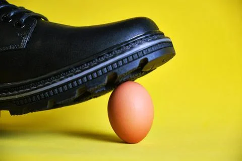Black boot on a yellow background stepped on a fragile chicken egg. Concept Stock Photos