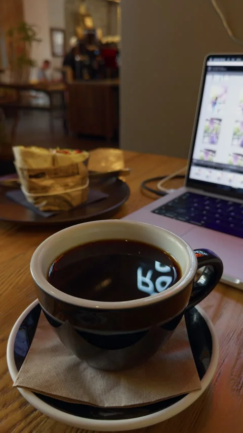 Black coffee cup beside laptop on wooden table in cafe with pastry and blurred Stock Footage 345606366