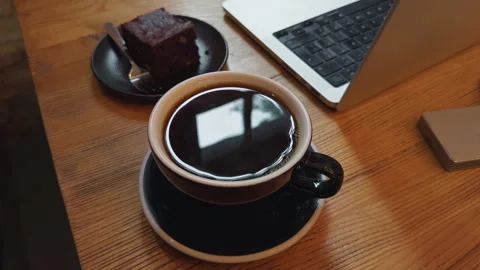 Black coffee in cup beside slice of chocolate cake and laptop on wooden desk Stock Footage 337648311