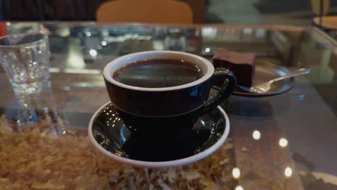 Black coffee cup on glass table with water glass, dessert square, and spoon in Stock Footage 346558796