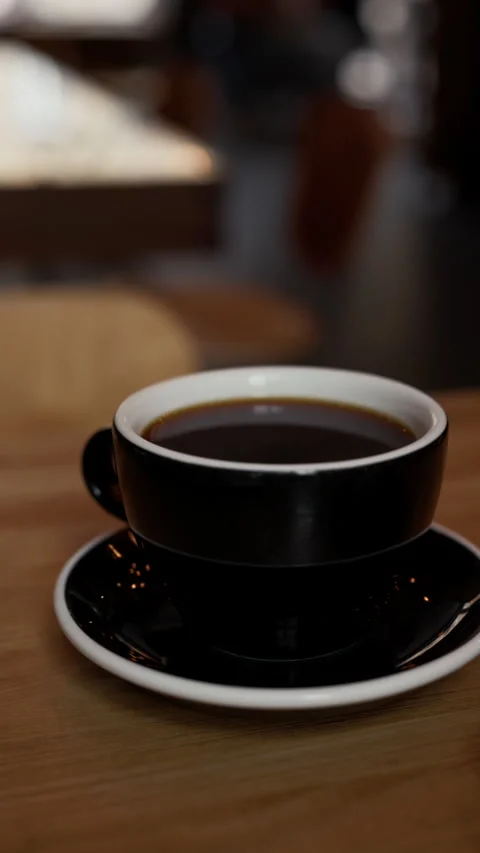 Black coffee cup on saucer with dark espresso on wooden table in softly blurred Stock Footage 345373339