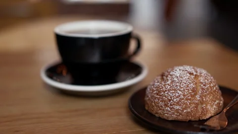Black coffee cup with sugar dusted pastry on saucer in warm cafe setting Stock Footage 345373464