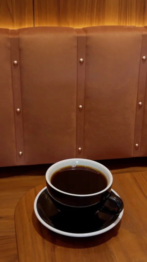 Black coffee cup on wooden table in cozy cafe booth with brown leather seating Stock Footage 345452482