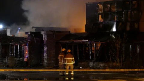 Black darkened building ablaze with pair of firemen standing in front Stock Footage 129986410