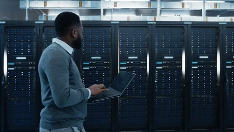 Black Data Center IT Technician Walking Through Server Rack Corridor with Laptop Stock Footage 100803655