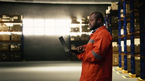 Black depot employee checking inventory data on laptop near racks Stock Footage 332908991