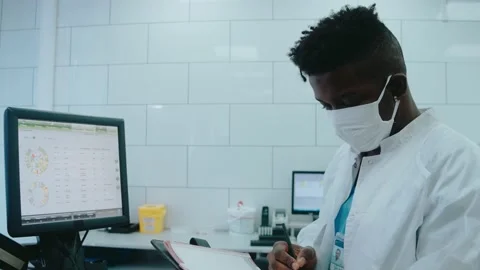 Black doctor writes information from computer in white lab wearing mask and gown Video stock 166666174