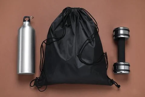 Black drawstring bag, thermo bottle and dumbbell on brown background, flat la Stock Photos