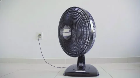 Black electric table fan plugged into a wall outlet in a white room Stock Footage 346644520