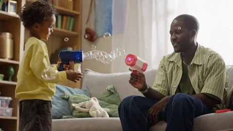 Black Father and Son Playing with Soap Bubble Guns in Living Room Stock Footage 345495343