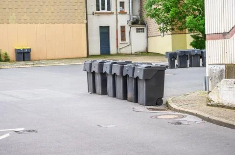 Black garbage bins lined along a residential street with buildings and green  Fotos de archivo