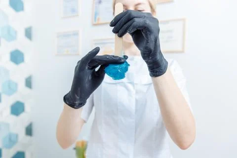 Black-gloved hands remove blue wax from a wooden spatula. Stock Photos