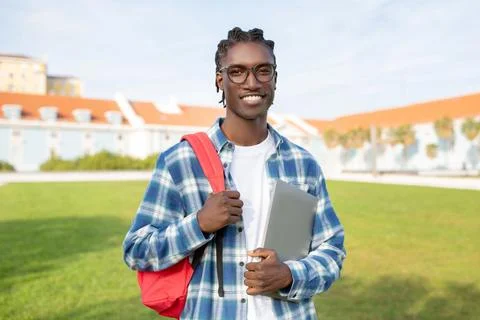 Black guy carrying backpack and laptop standing against campus backdrop Stock Photos
