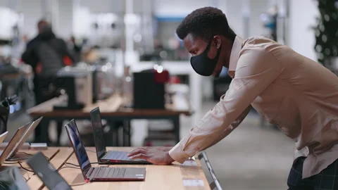 Black guy with face mask is choosing modern laptop for work in electronics shop Stock Footage 153075104